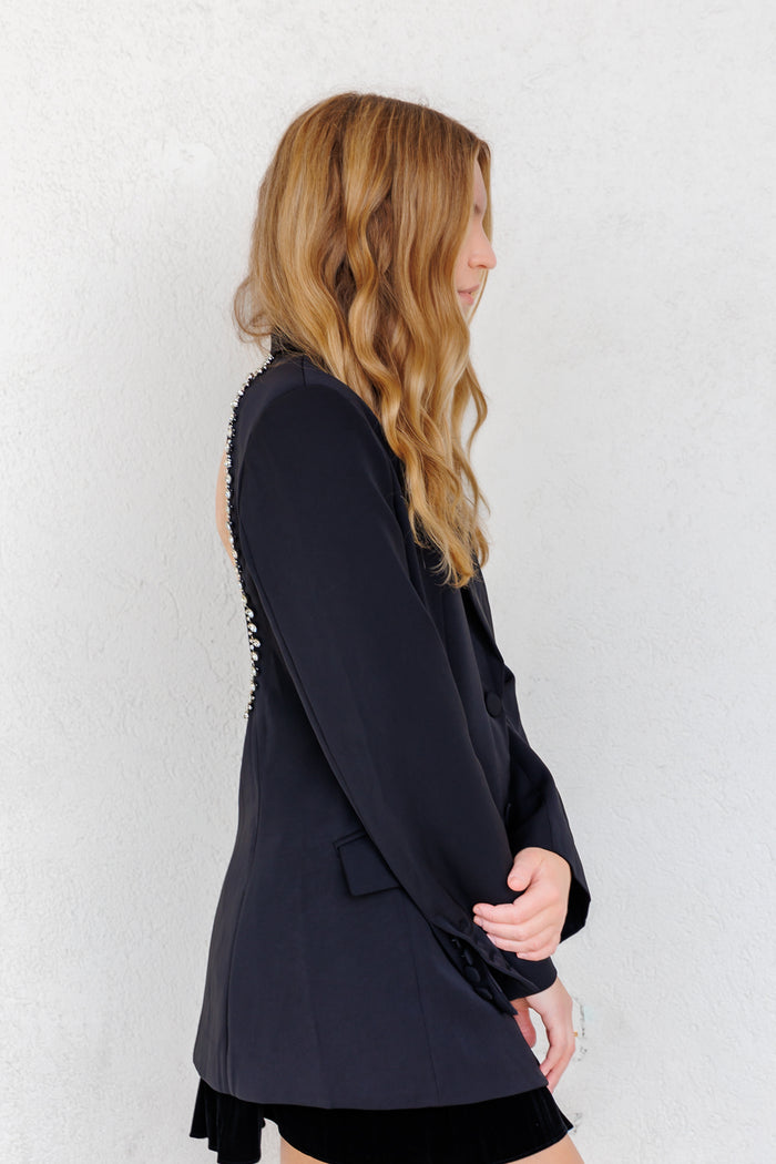 Black single-breasted blazer with pearl button detailing along the back seam, shown on model in profile view.
