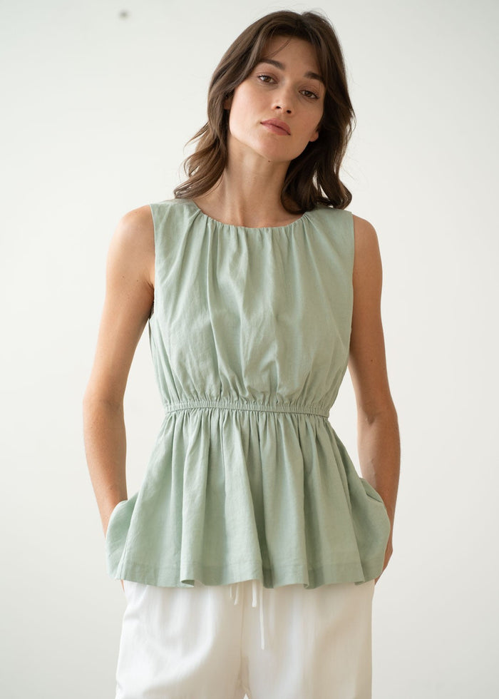 Sage green sleeveless peplum tank top with gathered waist and side pockets, paired with cream pants.