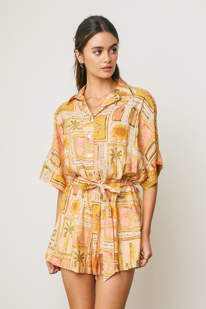 Woman wearing yellow and pink patterned short sleeve romper with sun and botanical print and tie waist detail.