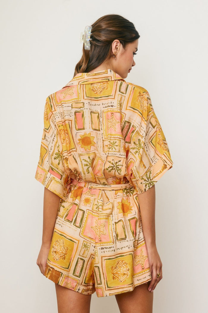 Woman wearing a short sleeve romper with a gold, pink, and cream botanical print featuring framed flowers and botanical sketc