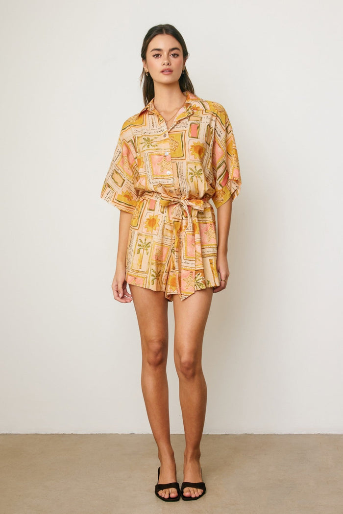 Woman wearing a short sleeve romper with a geometric and floral print in gold and pink tones, cinched at the waist.