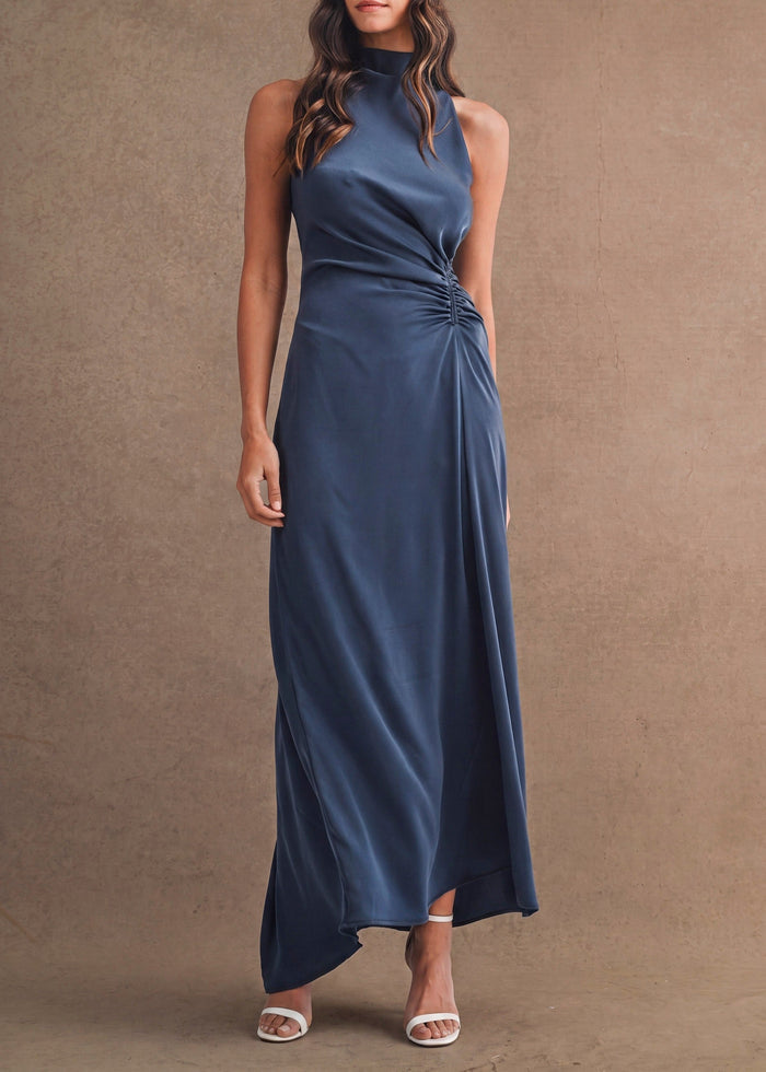 A woman modeling a sleeveless navy blue maxi dress with a high mock neck, side twist detail, and draped skirt, standing against a textured brown studio backdrop.