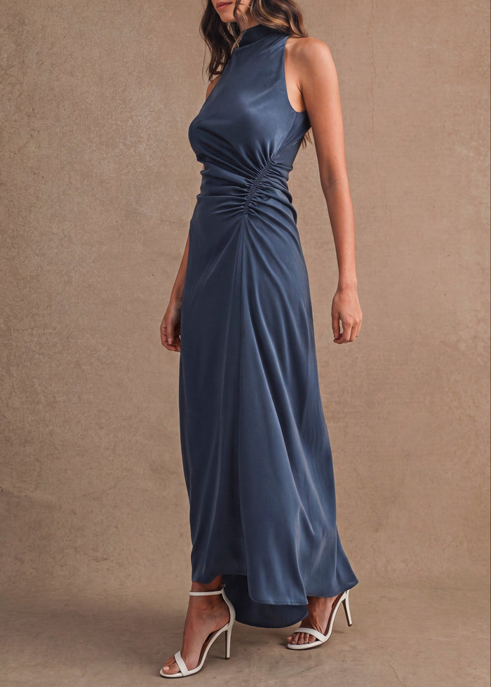 A woman modeling a sleeveless navy blue satin maxi dress with a gathered ruched waist, standing against a textured beige studio backdrop.