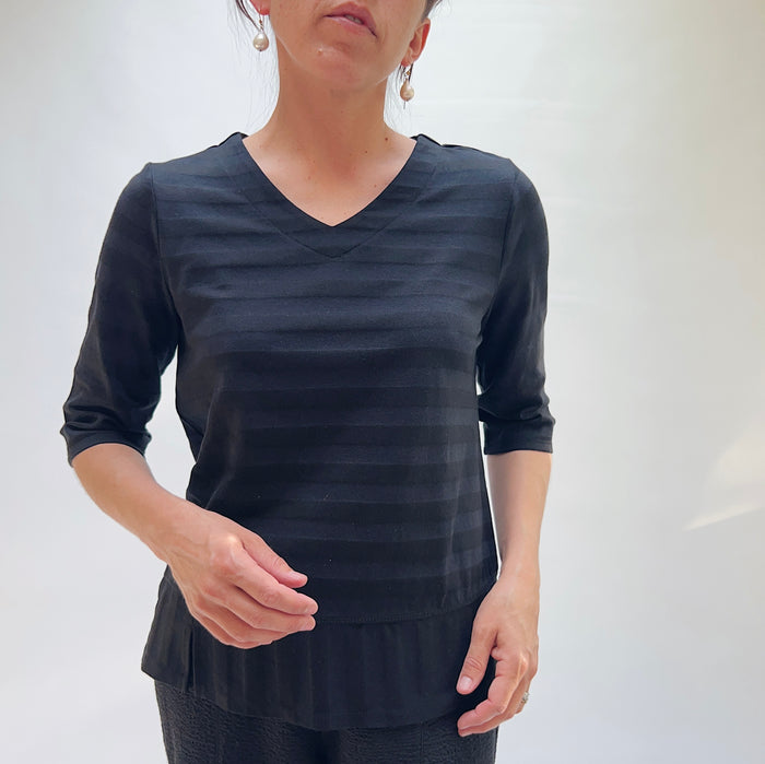 Close front view of model wearing Habitat black shadow stripe elbow sleeve top, showing V-neckline, tonal horizontal stripe texture, and layered hem with side slit