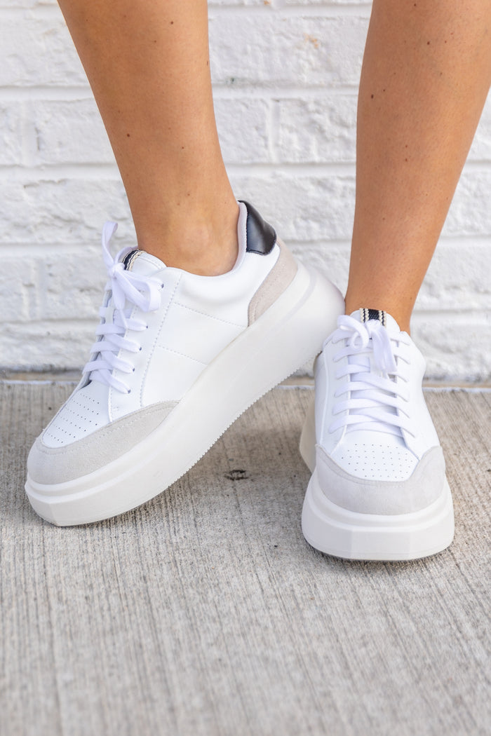 White lace-up sneakers with gray and navy accents on a chunky platform sole, worn with white socks.