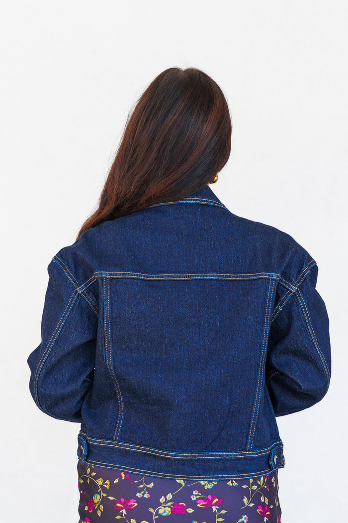 Dark blue denim jacket with contrast stitching, shown from the back on a model wearing a floral skirt.