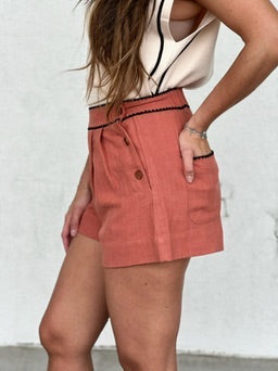 Terracotta textured pleated shorts with button waistband and side pockets, paired with cream sleeveless top.
