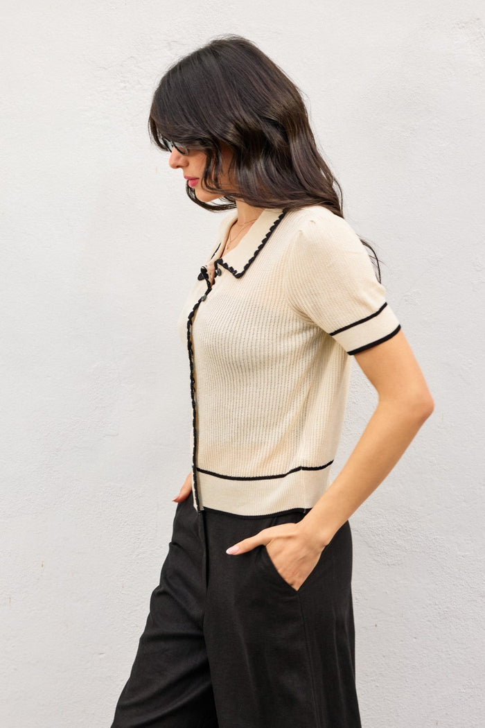 Cream ribbed knit top with black contrast trim, button front, and short sleeves worn with black pants.