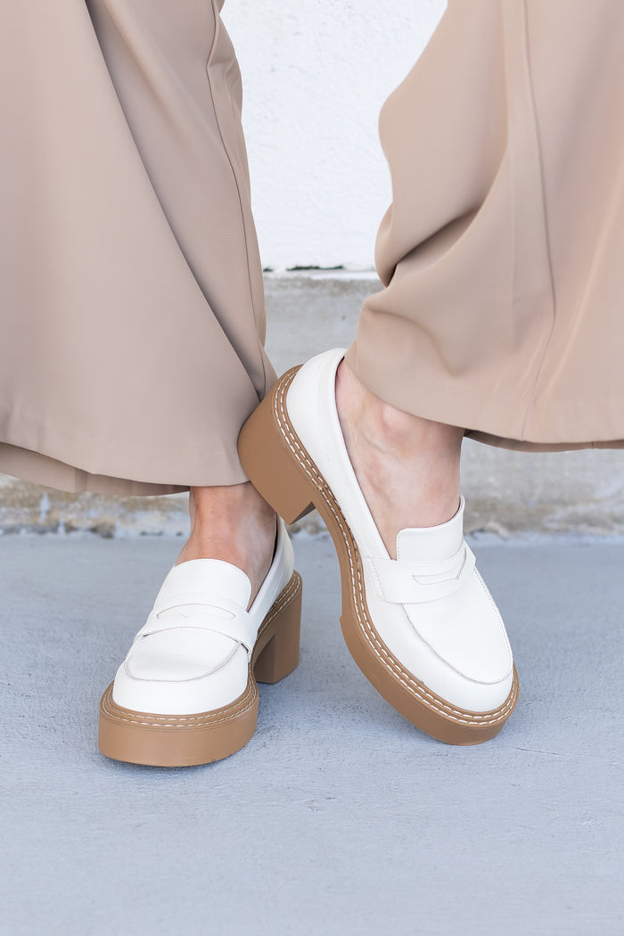 White leather loafers with tan chunky platform sole and stacked heel, worn with beige trousers.