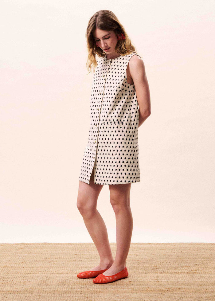 Cream sleeveless shift dress with small black diamond pattern and side slit, worn with red flat shoes.