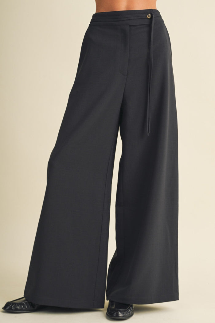 Black wide-leg trousers with a drawstring waistband, shown on a model from the waist down against a solid beige background.