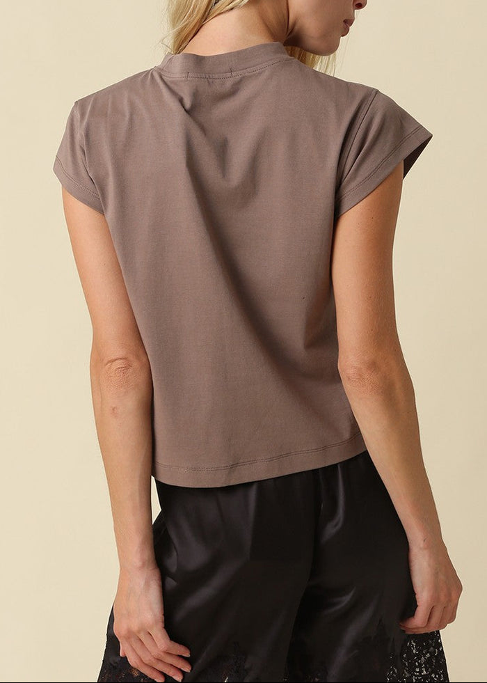 A woman shown from the back wearing a taupe short-sleeve crop top and black pants, photographed against a solid light beige background.