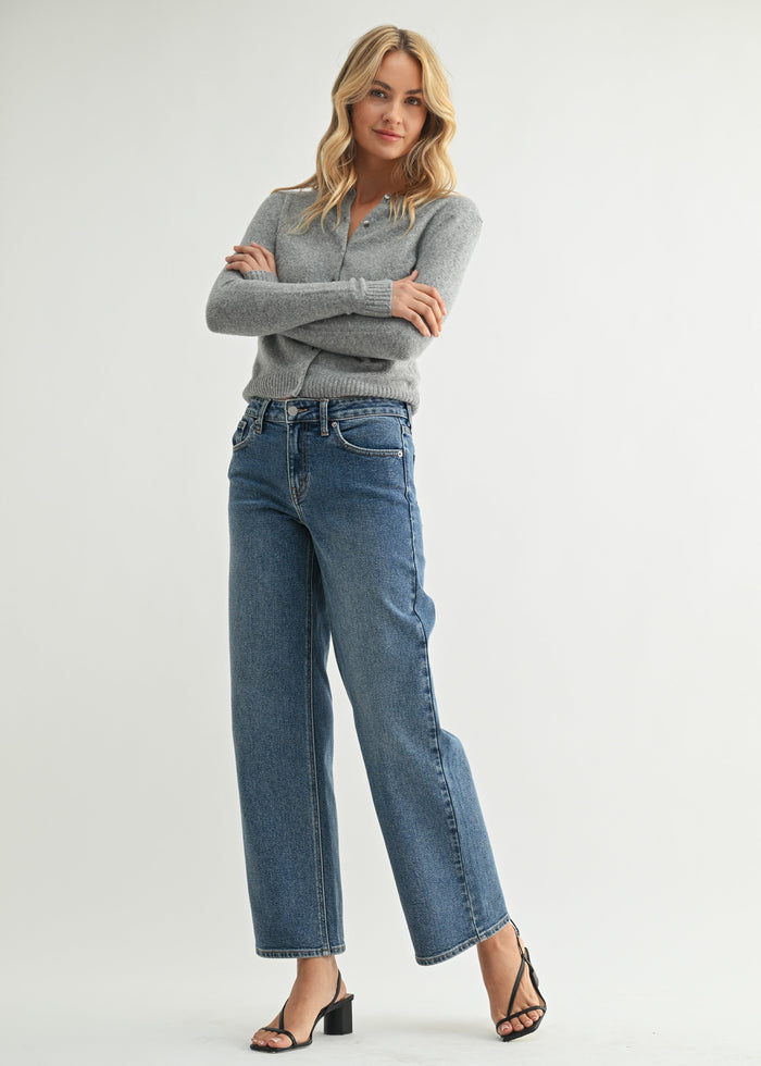 Woman wearing Victoria mid rise jeans in medium blue denim with straight leg, paired with gray sweater and black sandals.
