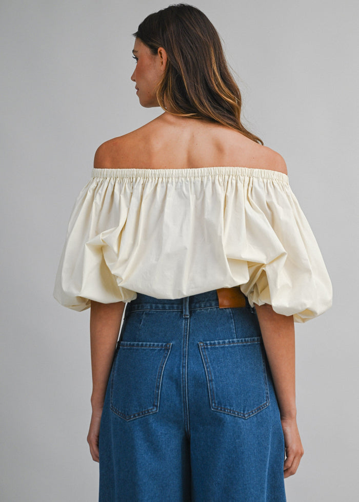 Cream off-shoulder top with gathered neckline and puffed sleeves worn with blue denim jeans, back view.