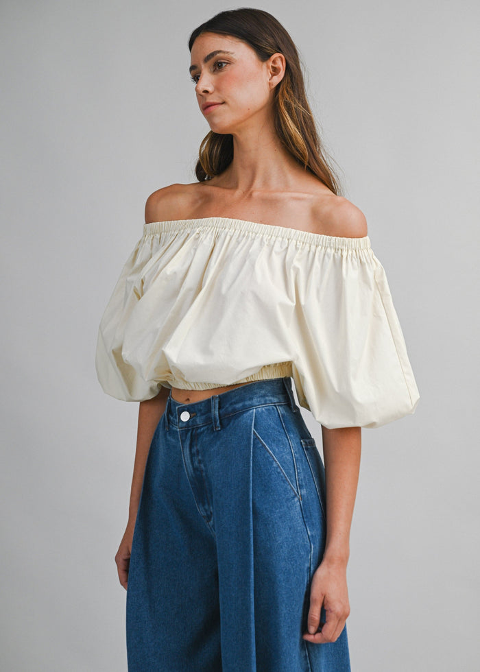 Cream off-the-shoulder top with puffed sleeves and elastic waistband, worn with dark blue denim jeans.